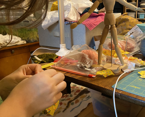 Child's hands sewing, with materials and small mannequin in the background